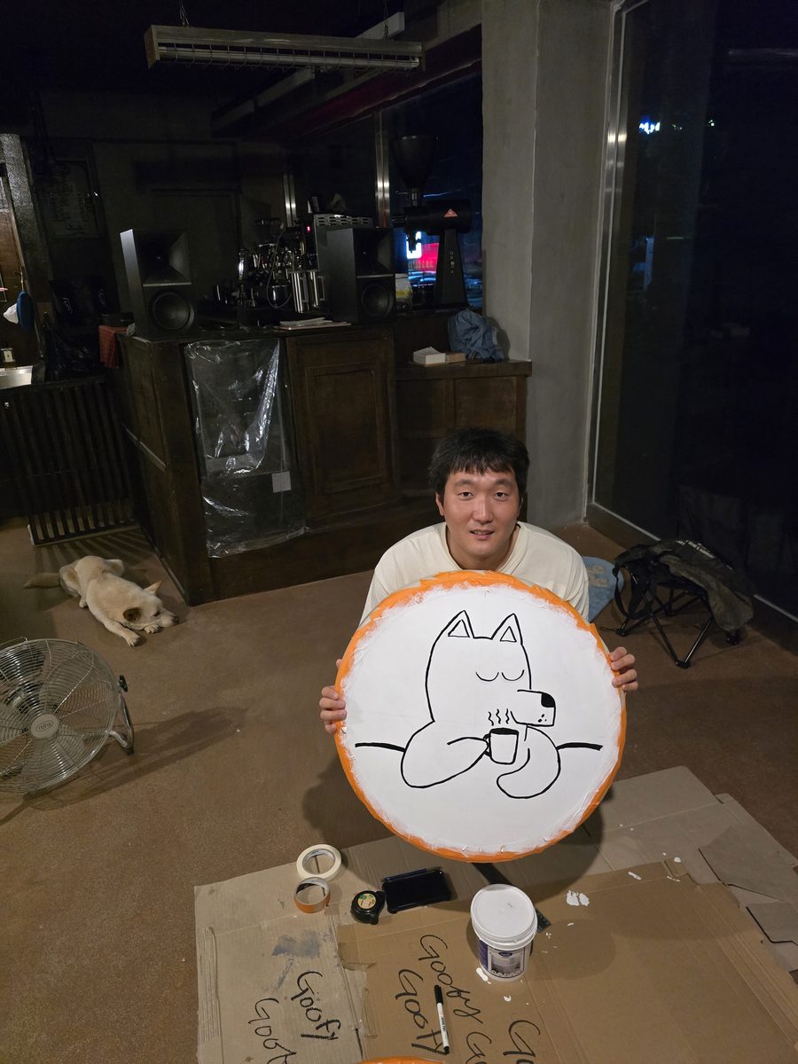 밤에 가게 바닥에 앉아 직접 그린 뭉개 간판을 들고 있는 사장, 뒤에 엎드린 뭉개
